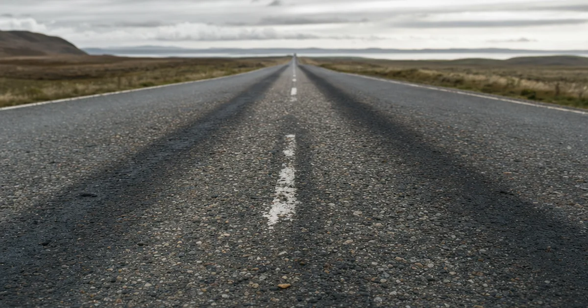 A worn asphalt road stretching to the horizon with faded centre-line markings, darker tyre-polished wheel paths, and a rough gritty surface